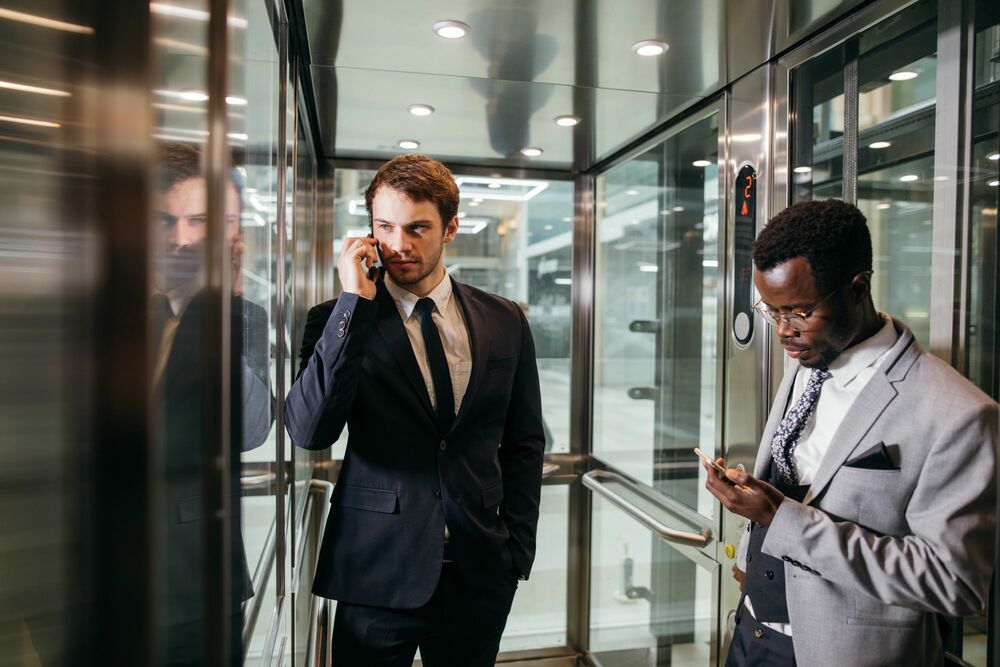 dois homens de terno posicionados como se analisassem algo, um fala ao telefone enquanto outro olha para o celular