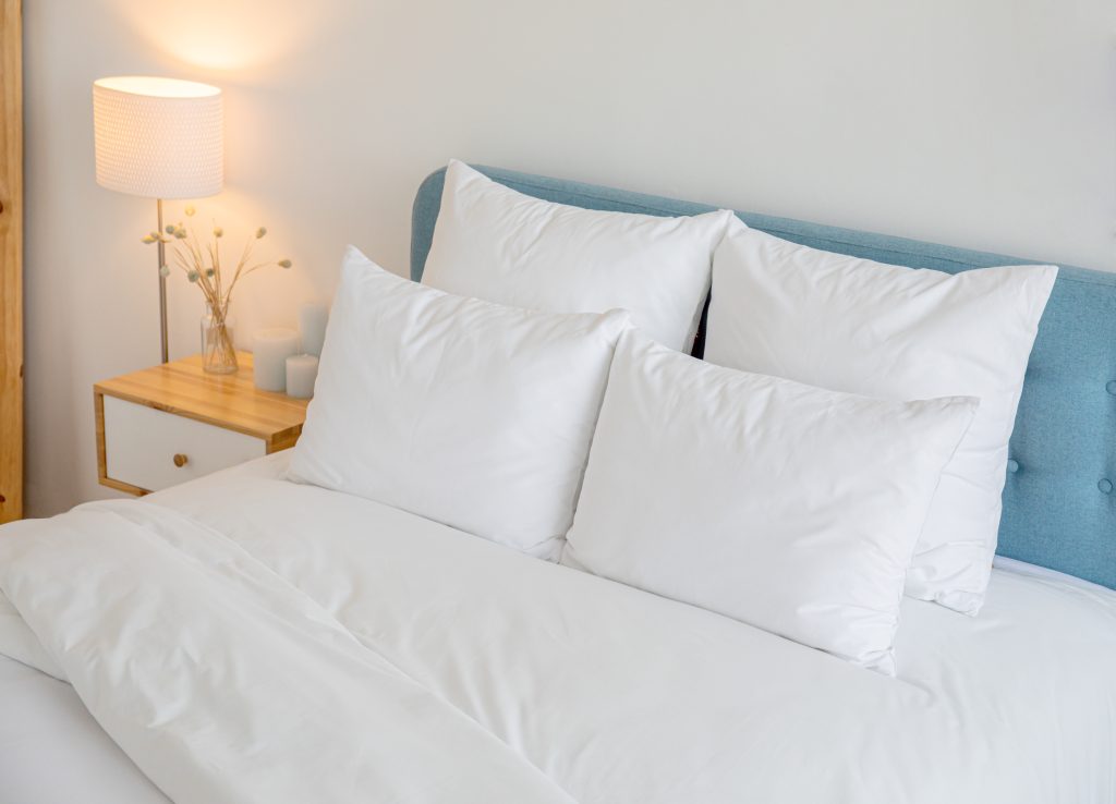 cama posta com travesseiros da cor branca, cabeceira em azul e detalhes amadeirados do móvel lateral.